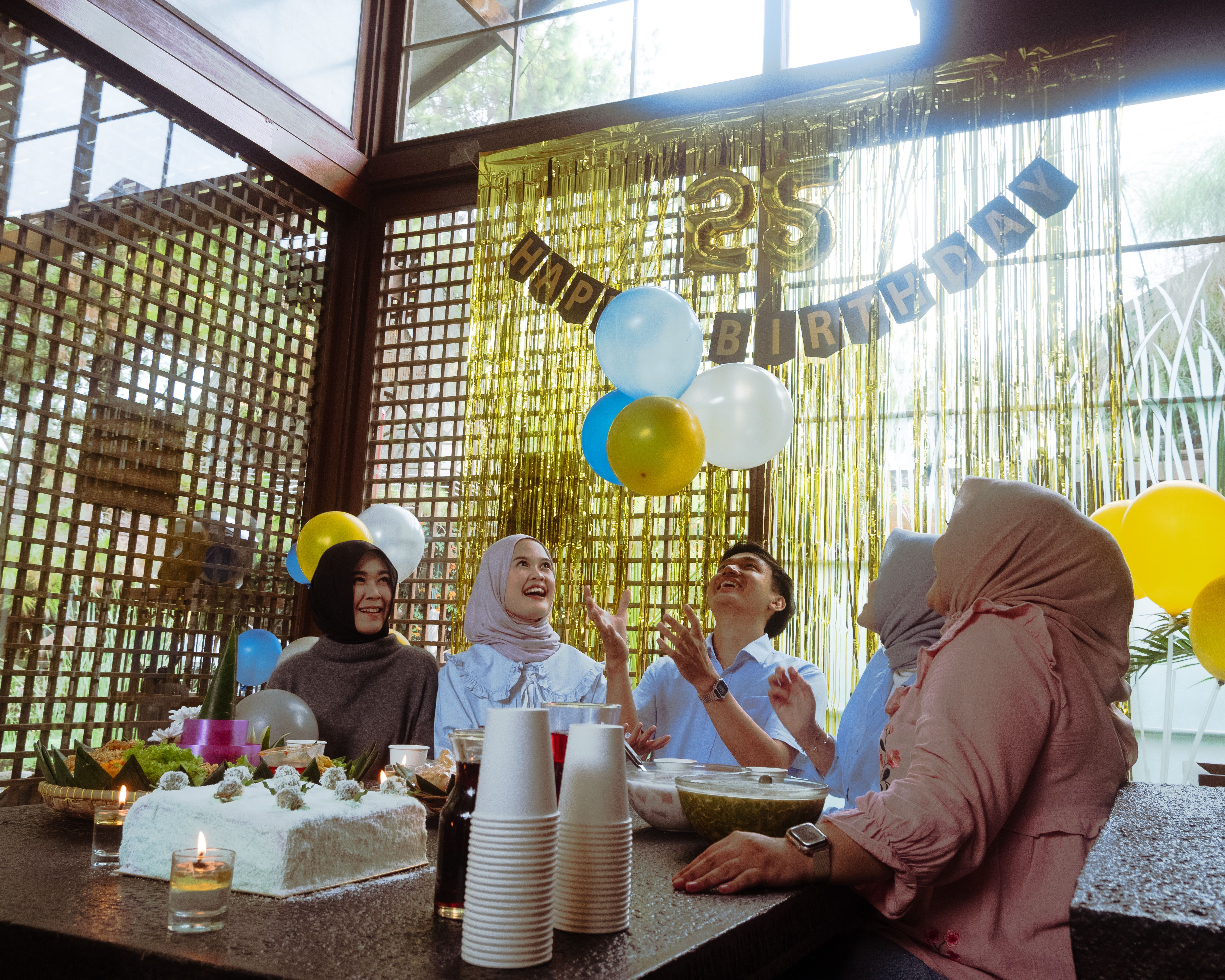 A lively indoor birthday party with friends, balloons, and cake in Jawa Barat, Indonesia.