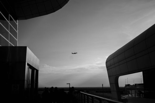 Free Black and white photo of an airplane flying above a modern airport terminal at sunset. Stock Photo