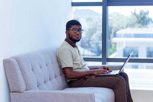 Young adult man working on a laptop while sitting comfortably on a sofa by a window.
