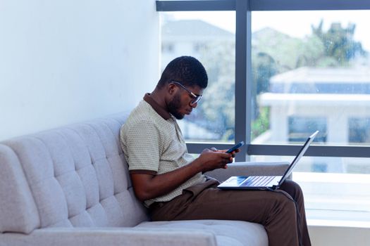 Man multitasking with laptop and smartphone indoors, showcasing remote work lifestyle.