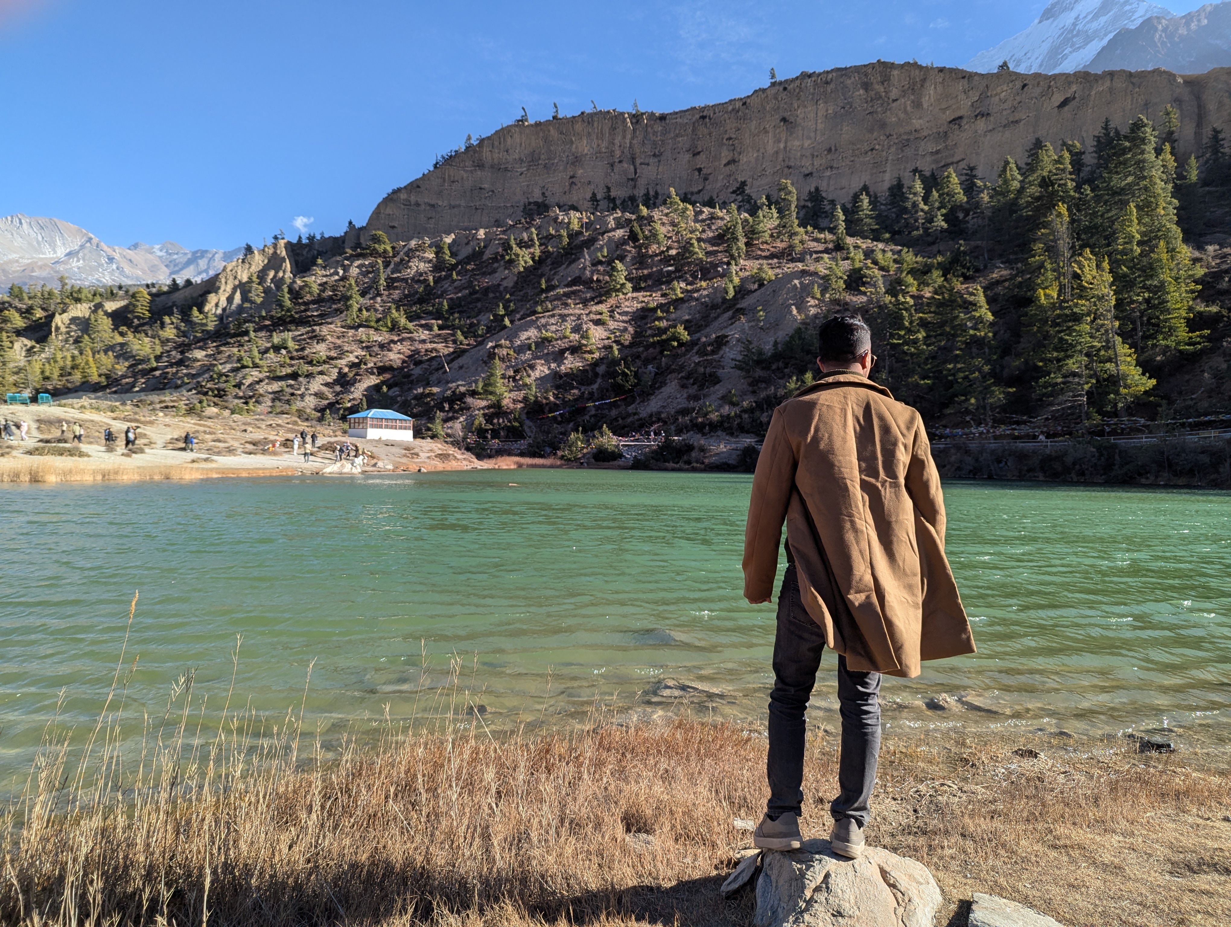 A man stands by a turquoise lake, gazing at the mountains and rocky landscape under a clear autumn sky.
