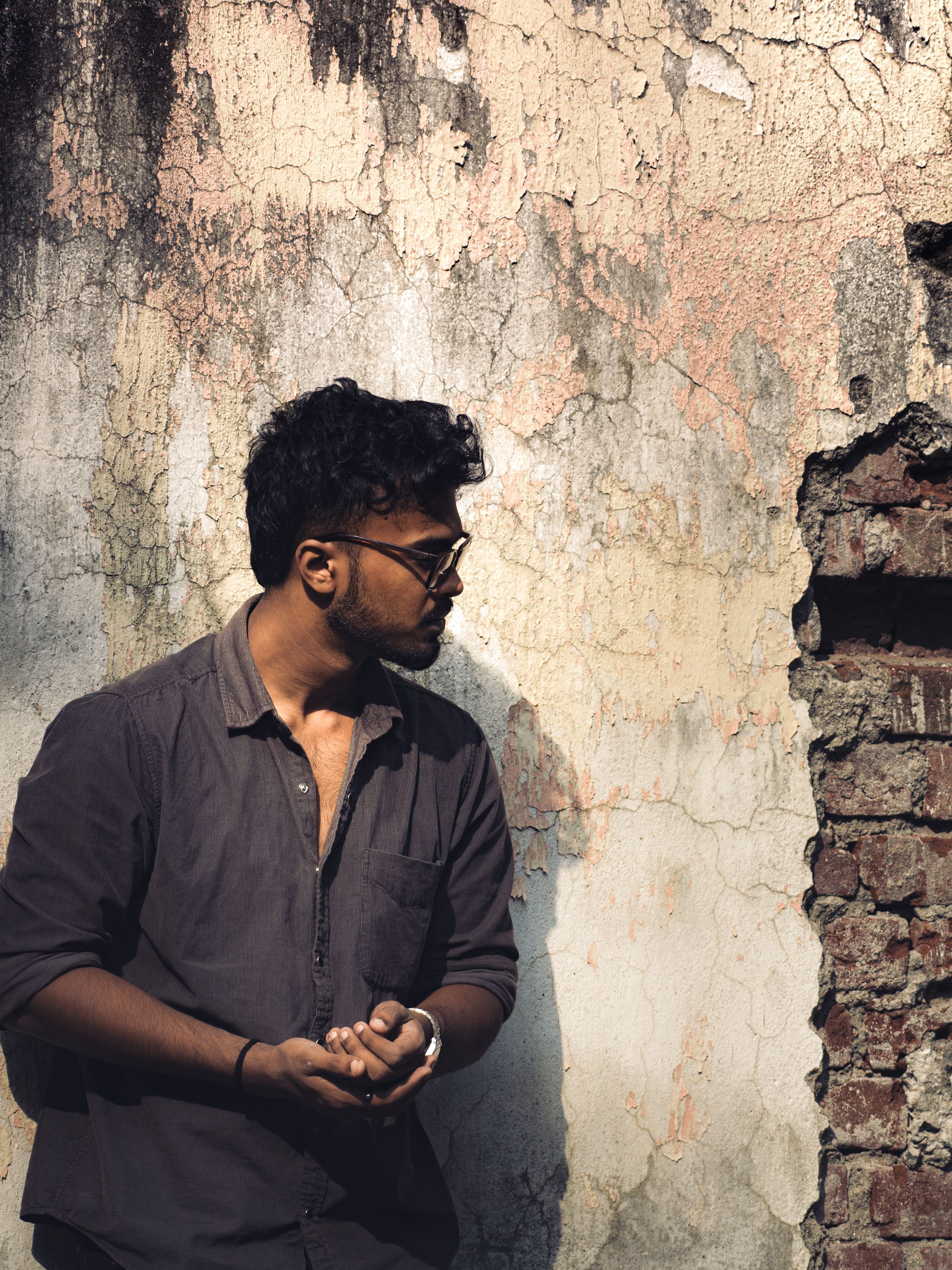 Young man standing against a rustic, weathered wall with a thoughtful expression in natural light.