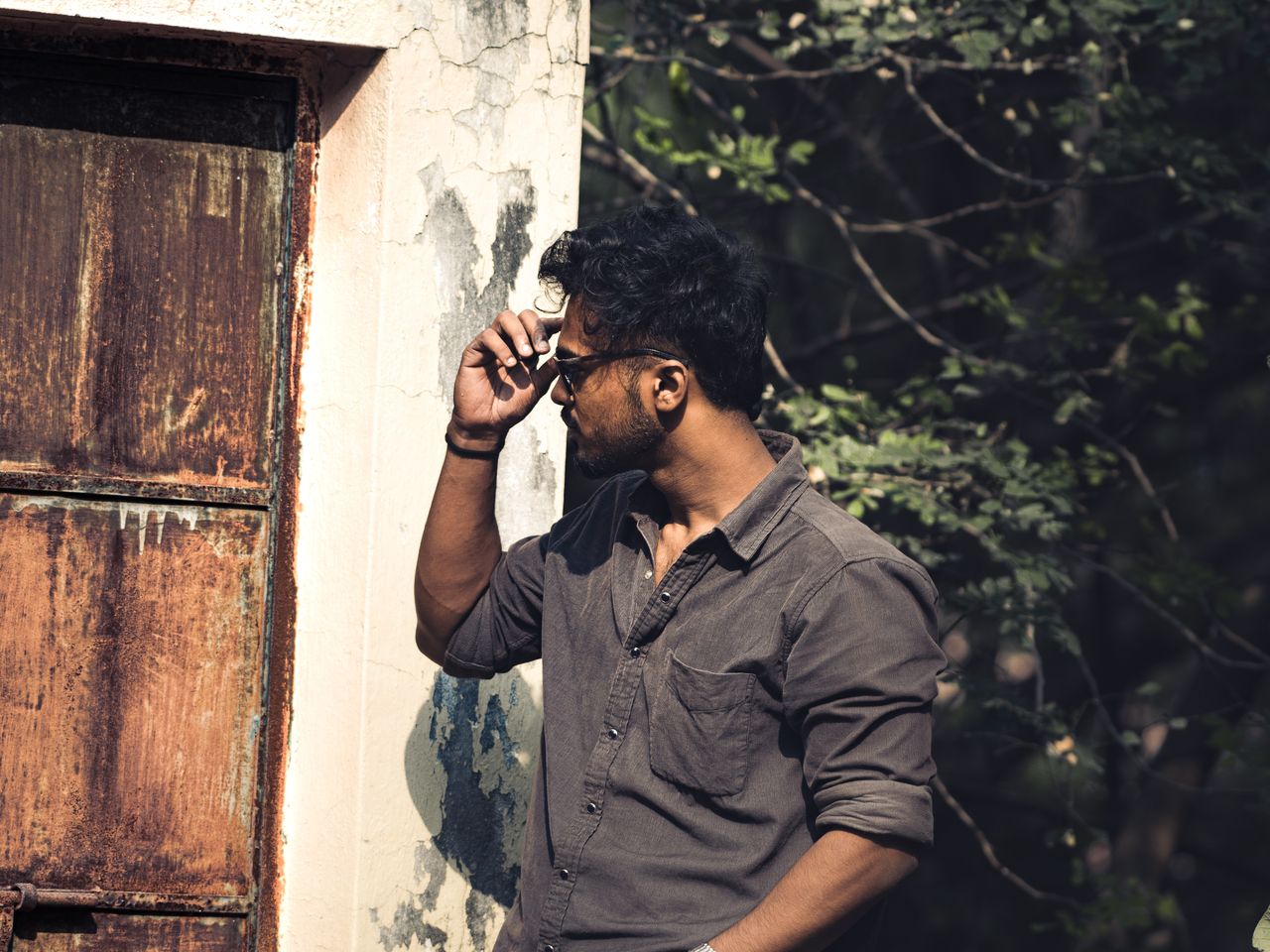 a man adjusting his sunglasses stands stylishly by a rusty door surrou