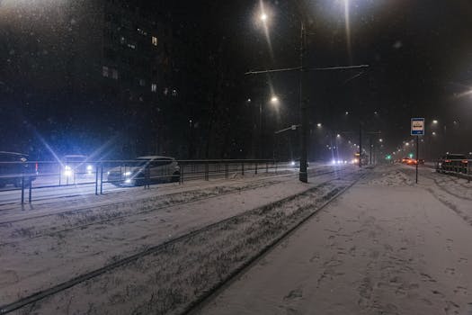 Night view of snowy street with cars and tram tracks under streetlights during snowfall.