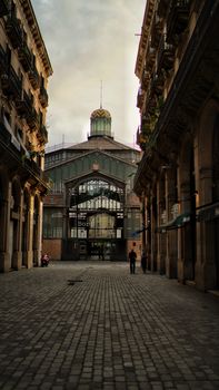 A quiet cobblestone street leading to a grand market building in Barcelona, Spain.