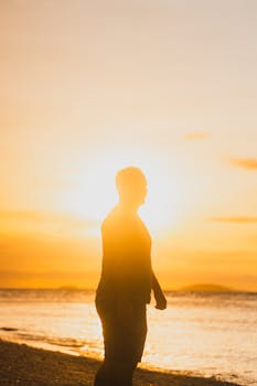 Silhouette of a person standing by the sea during a vivid sunset, creating a peaceful beach atmosphere.