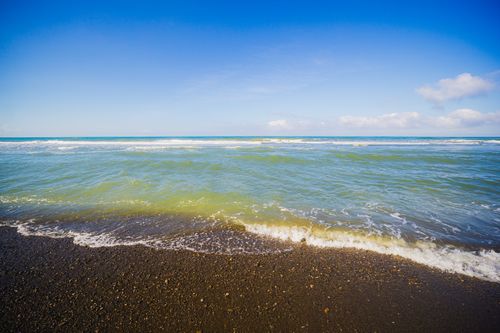 Free A serene beach scene with gentle waves, clear blue sky, and dark sand under sunlight. Stock Photo