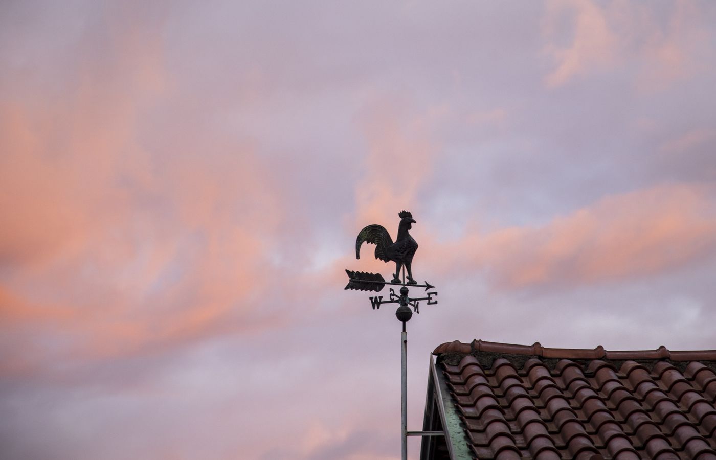 Silhouette of a weather vane against a colorful sunset sky over a tiled rooftop, capturing rustic charm.
