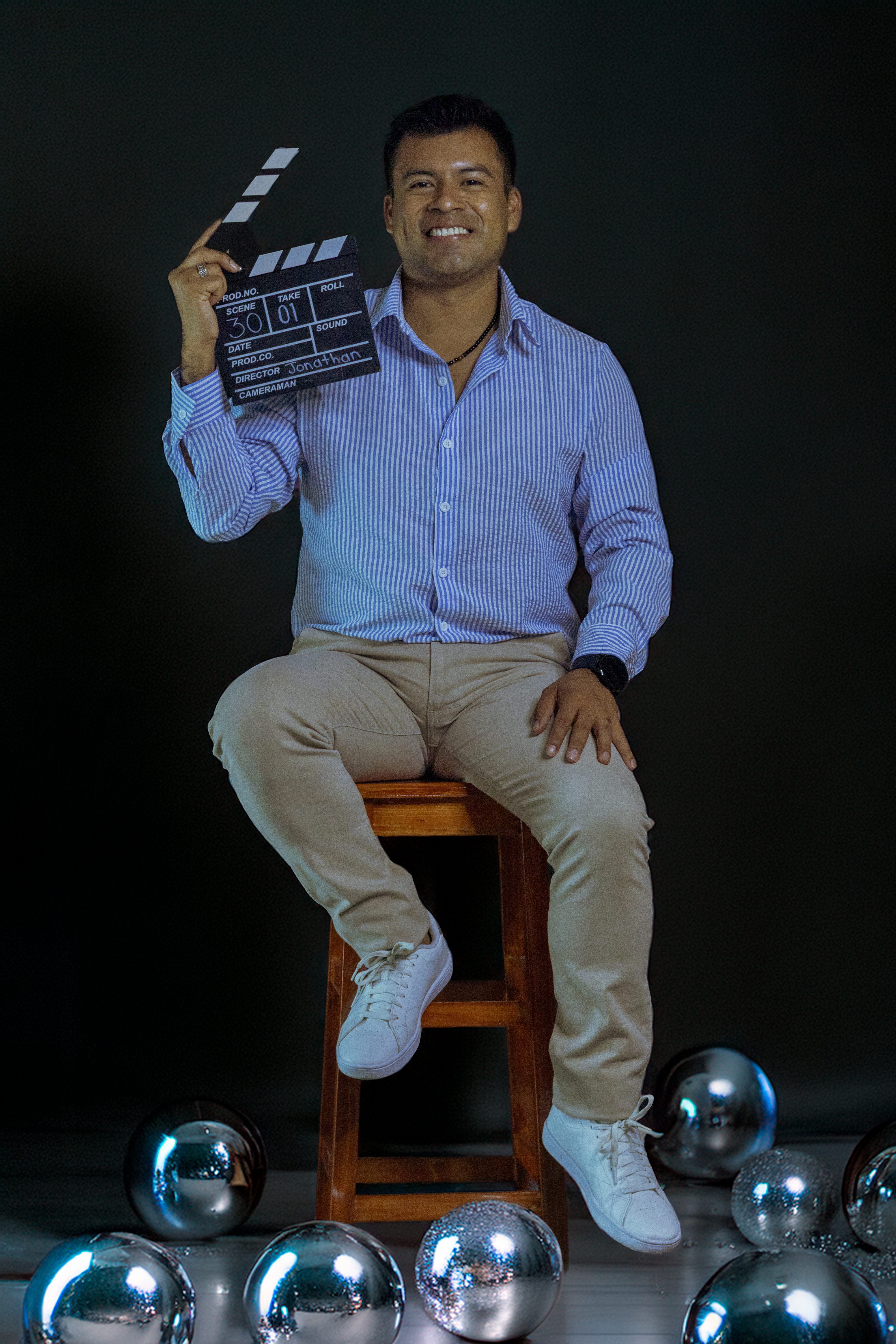 Smiling man holding a clapperboard sitting among reflective spheres in a studio.