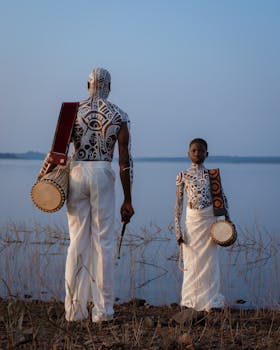 Two traditional drummers with symbolic body paint stand by the Lagos Lagoon at sunset, showcasing Nigerian culture.