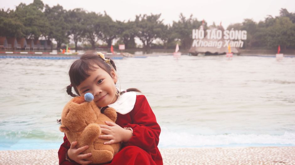 A young girl in a red dress hugs her teddy bear by the water at Khoang Xanh Park.