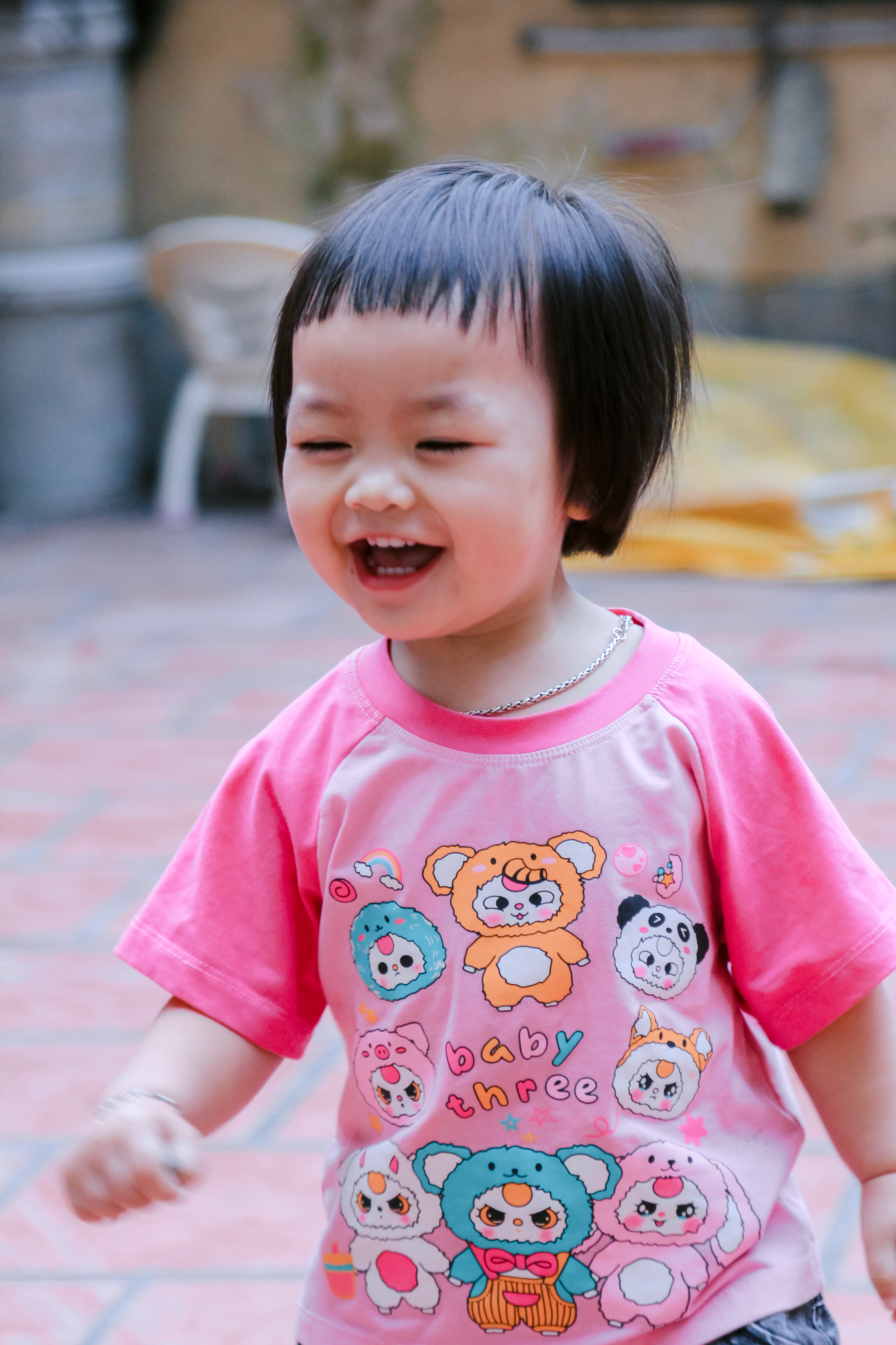 Adorable toddler laughing, wearing a pink shirt with cute cartoon designs, outdoors.