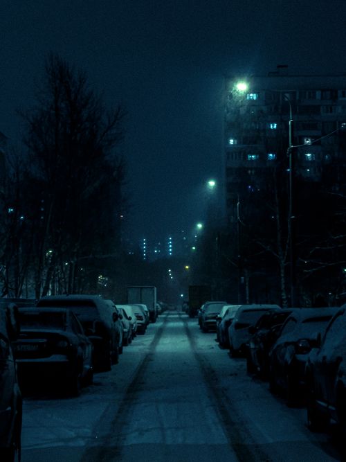 Free A quiet, snow-covered urban street at night, lined with parked cars and illuminated by streetlights. Stock Photo