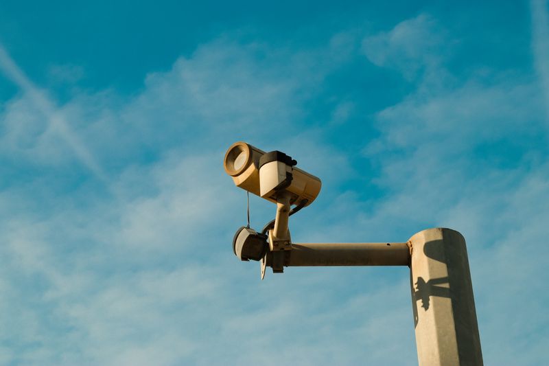 A surveillance camera mounted outdoors against a bright blue sky, symbolizing security and monitoring.
