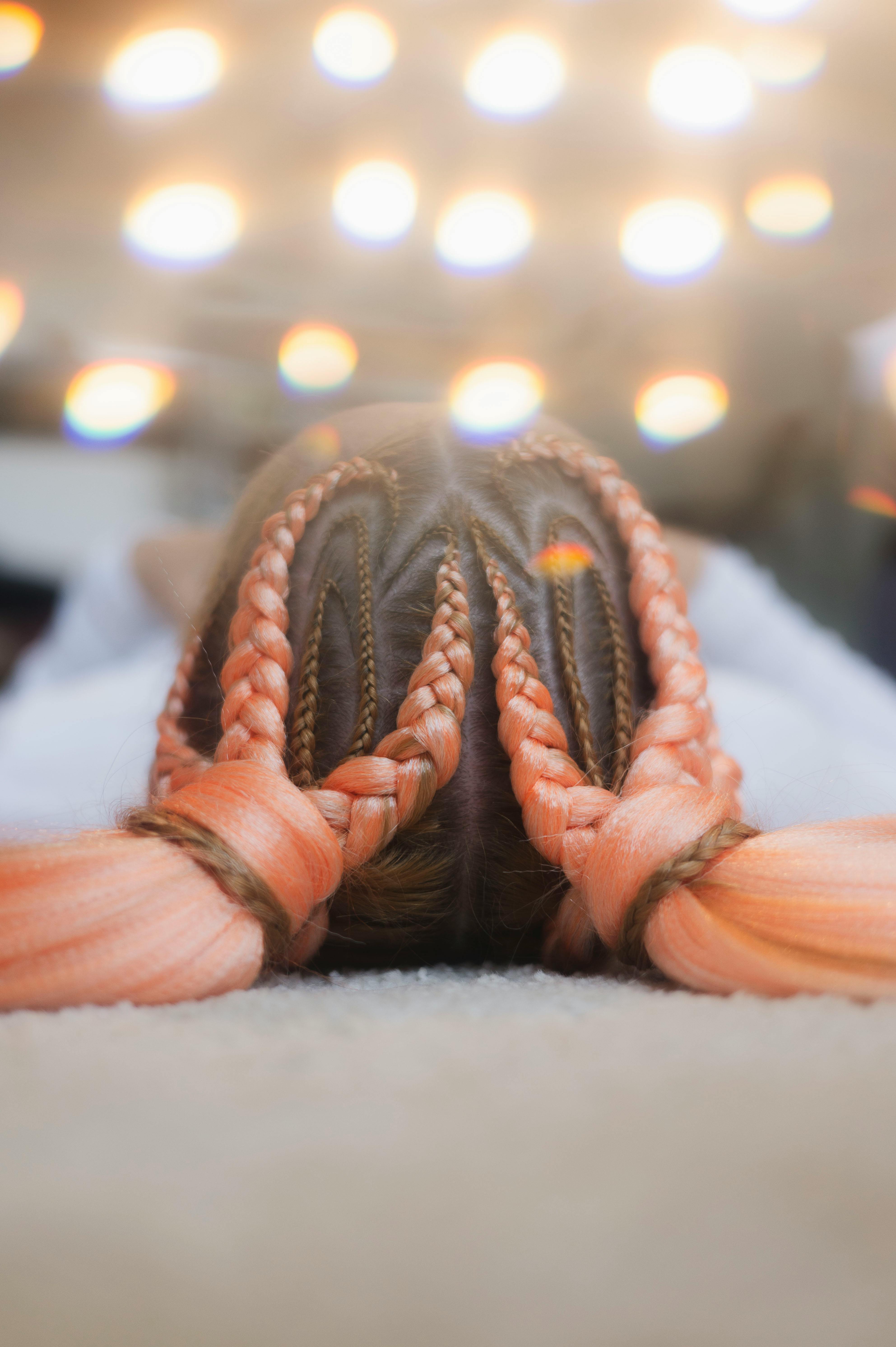 A close-up of a vibrant braided hairstyle featuring pink and orange hues, with a soft bokeh light effect.