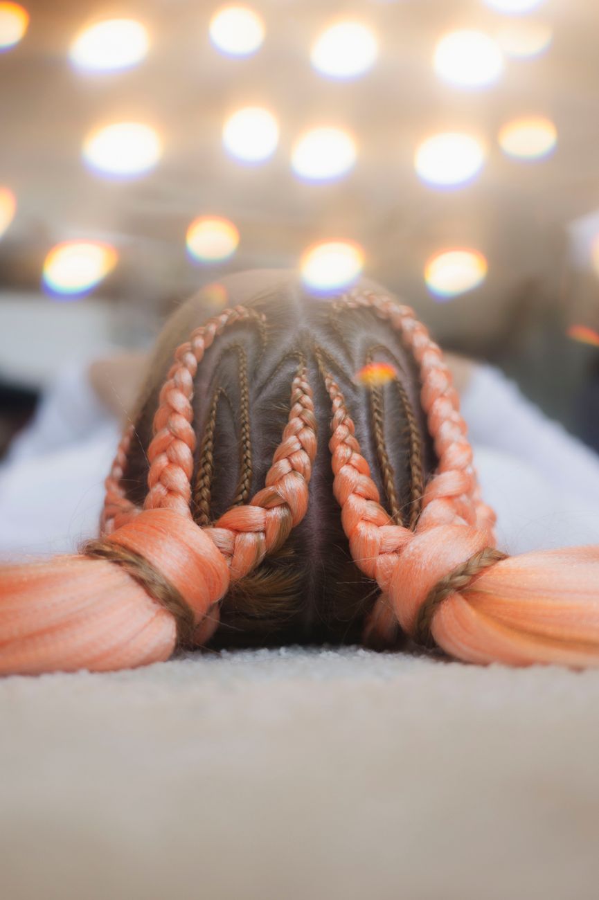 A close-up of a vibrant braided hairstyle featuring pink and orange hues, with a soft bokeh light effect.