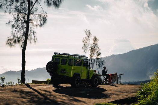 A person sits beside a green Jeep exploring the stunning mountain landscape of East Java, Indonesia.