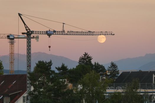 A construction crane silhouetted against a full moon, captured during dusk with mountain backdrop.