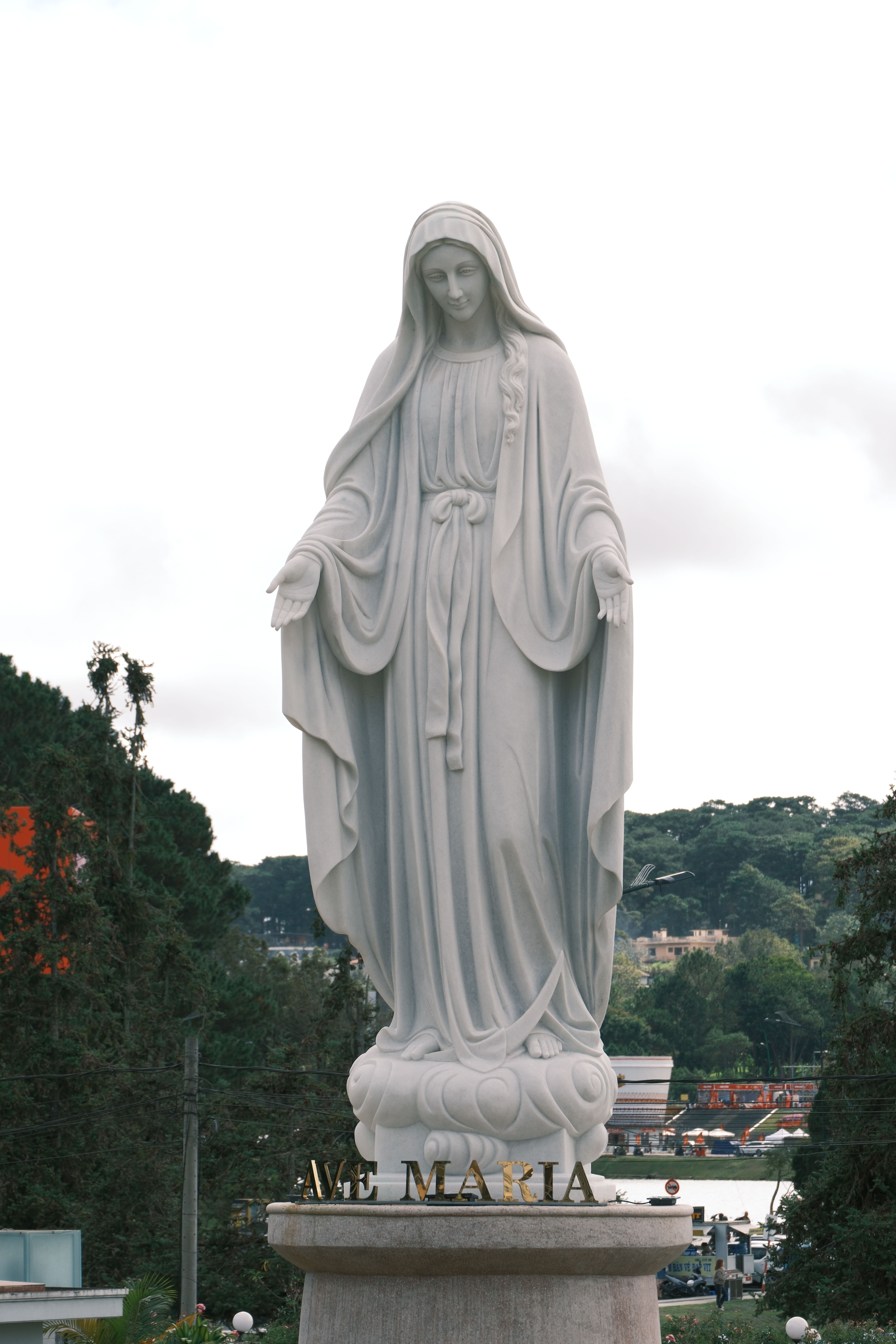 White statue of Virgin Mary with outstretched hands in an outdoor garden.