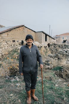 Older shepherd standing outdoors near a stone barn with sheep in the background.
