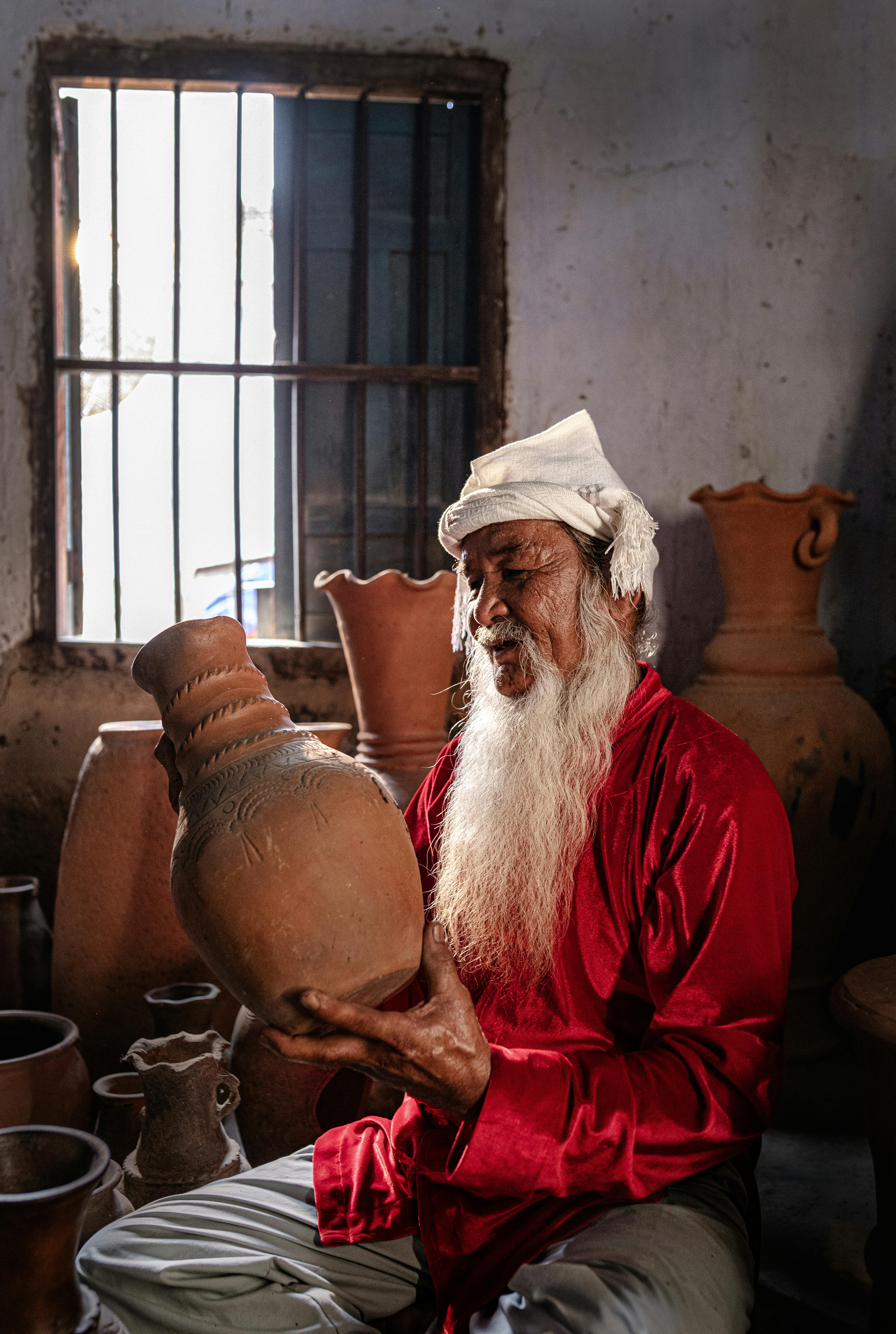 An elderly potter in vivid attire inspects a handcrafted clay jug in a rustic workshop.