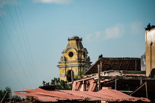 Free A historic yellow clock tower stands tall with pigeons perched on rooftops, against a bright sky. Stock Photo