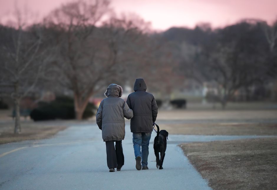 Couple walking dog in park at twilight, winter coats on, calm atmosphere.