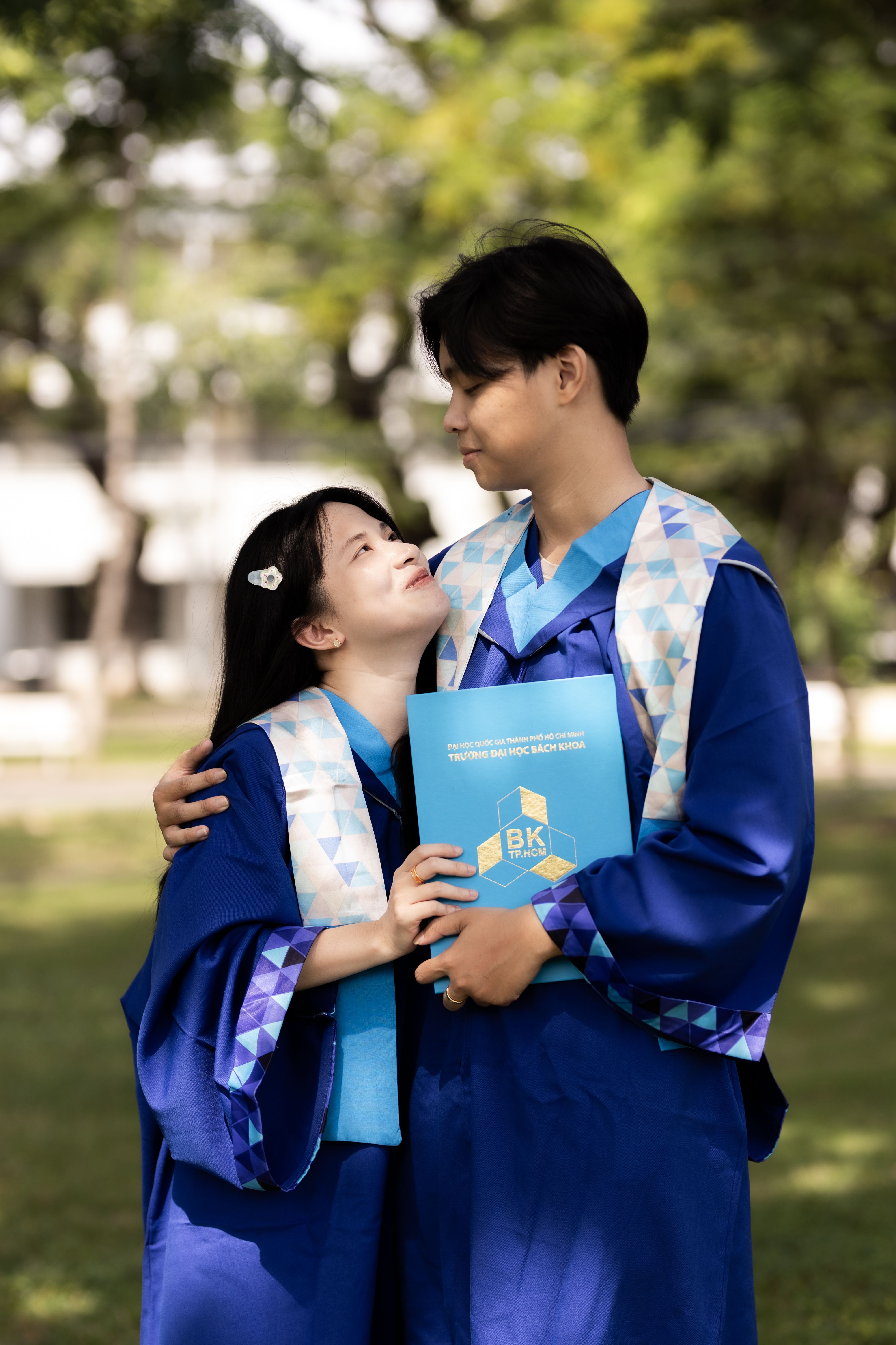 Two graduates in blue robes celebrate their achievement outdoors in Ho Chi Minh City, Vietnam.