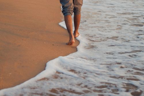 Free A person walks barefoot along a sandy beach with gentle waves at sunrise, capturing a peaceful moment. Stock Photo
