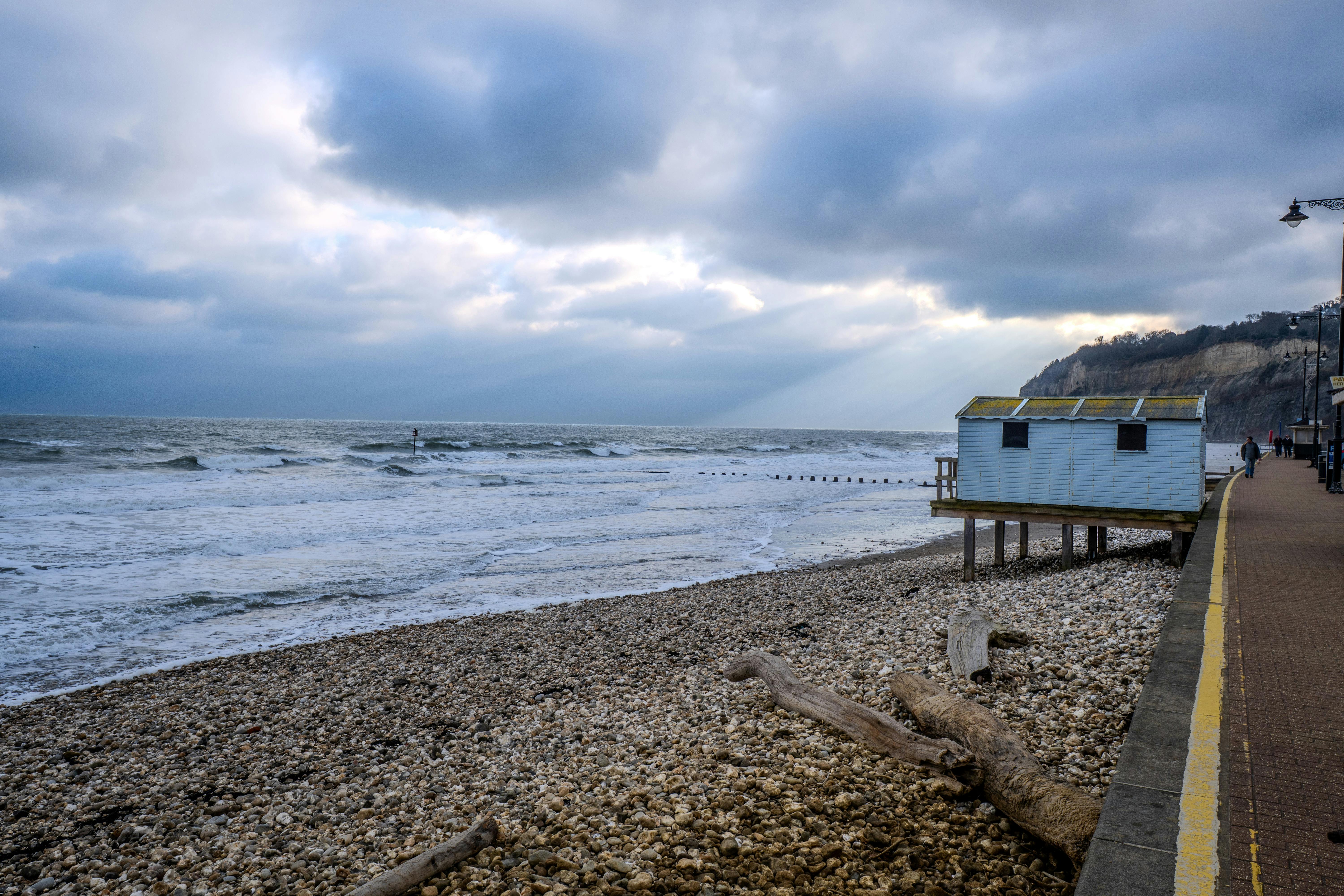 Tranquil seaside scene with a beach hut, pebbled shore, and waves under a cloudy sky.