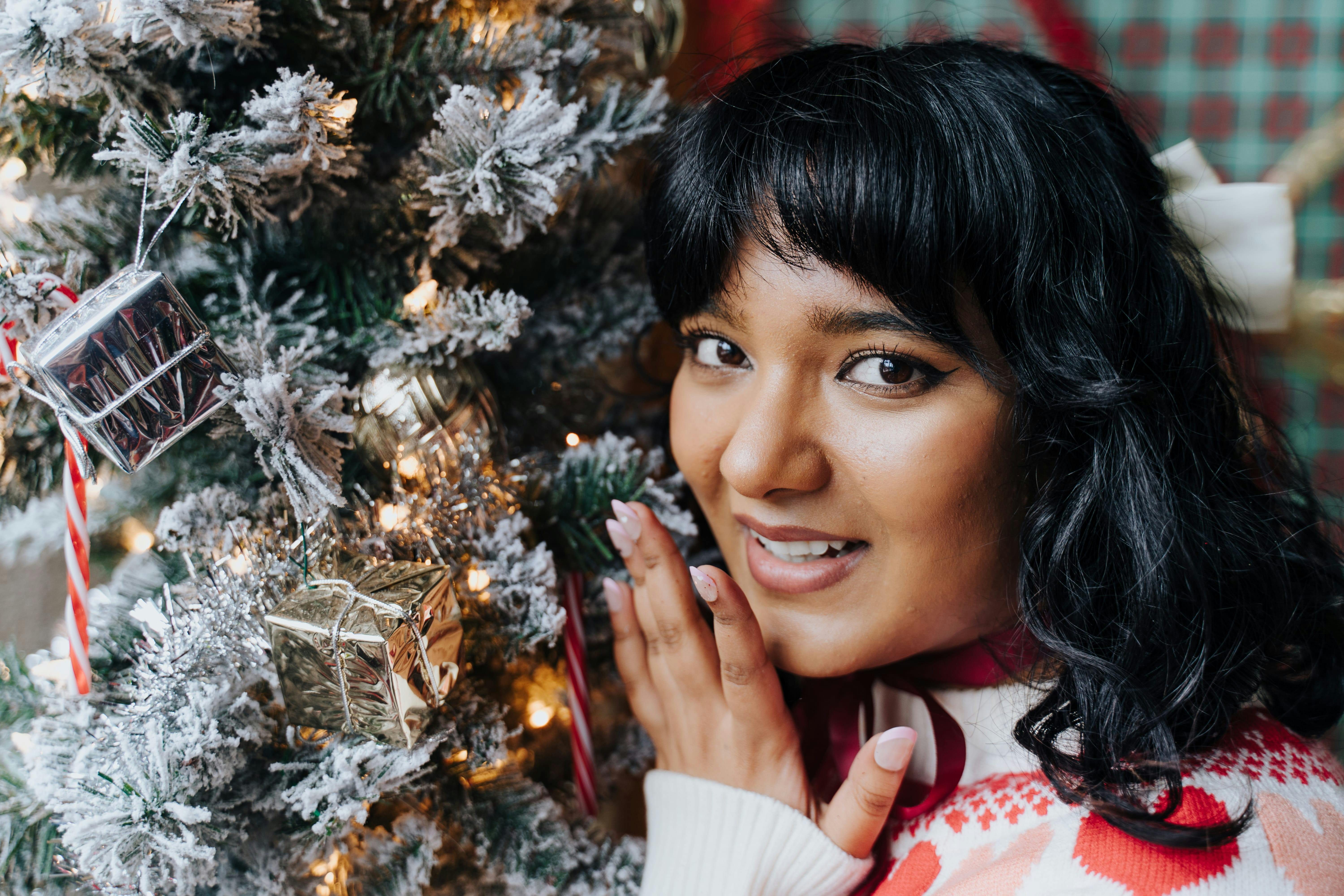 Zadarmo Radostná mladá žena pózuje vedľa ozdobeného vianočného stromčeka v interiéri. Fotka z fotobanky