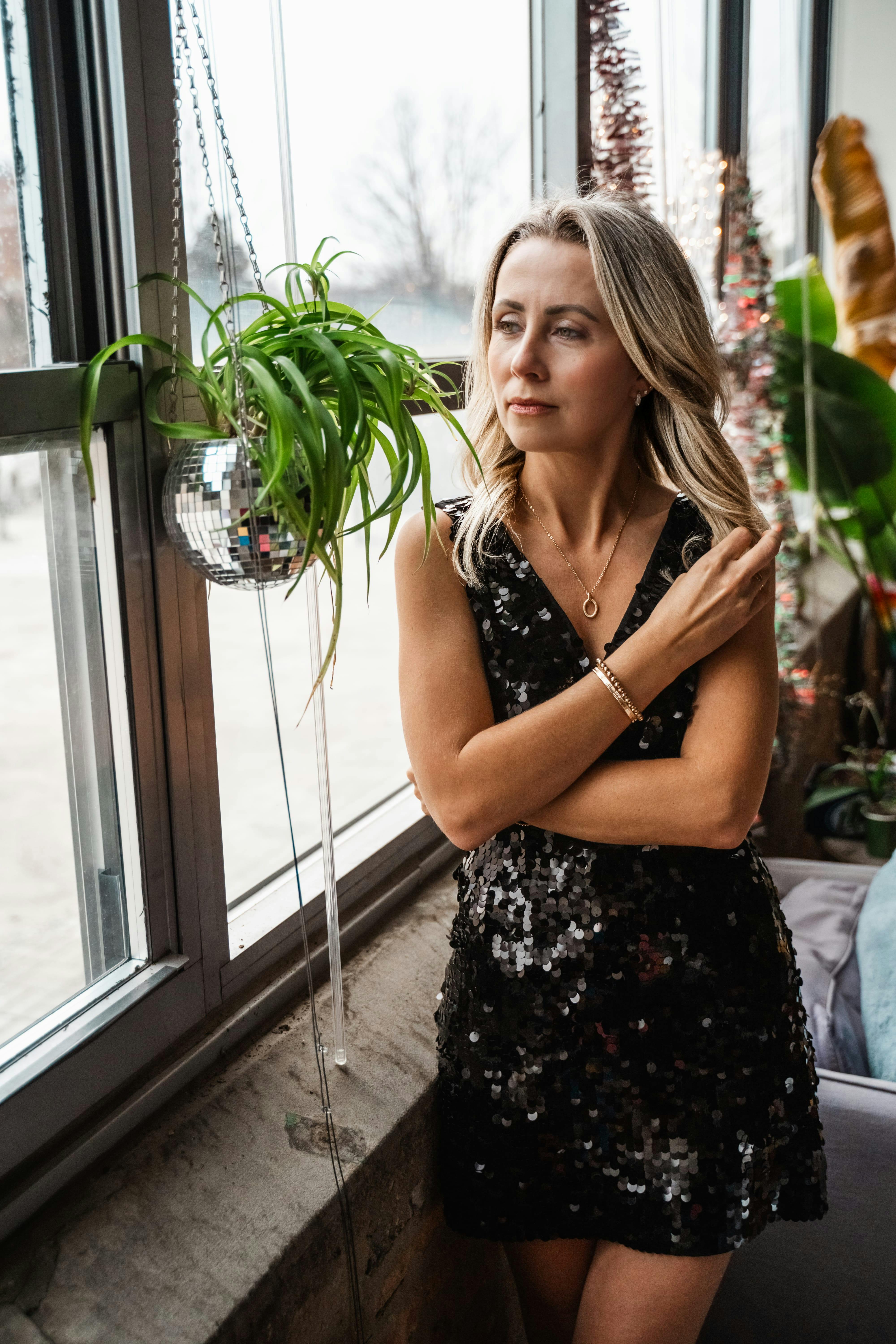 Zadarmo Štýlová žena v čiernych flitrovaných šatách stojaca pri okne, obklopená rastlinami a dekoráciami. Fotka z fotobanky