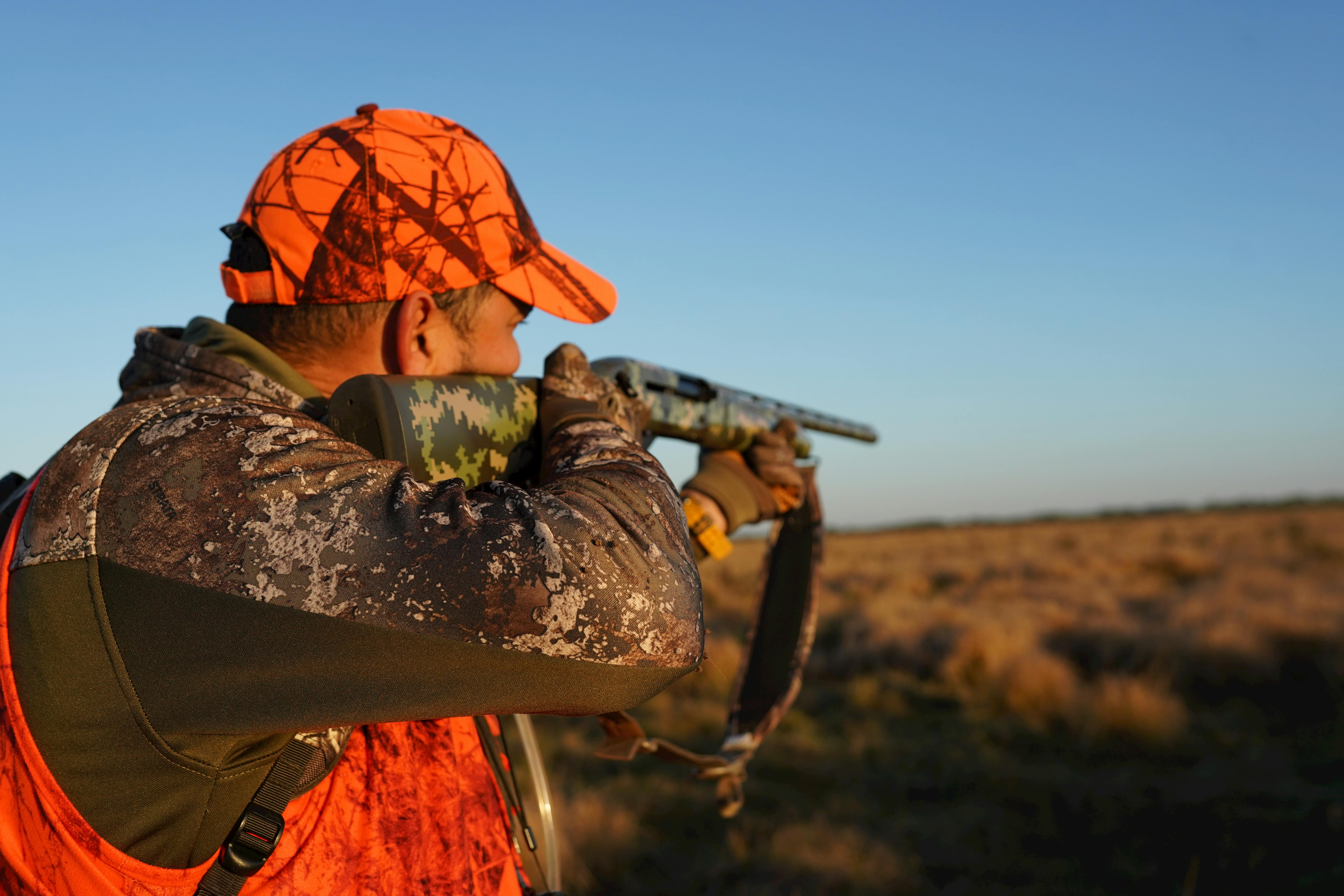 An adult man in camo and orange gear aims his rifle in a Florida field.