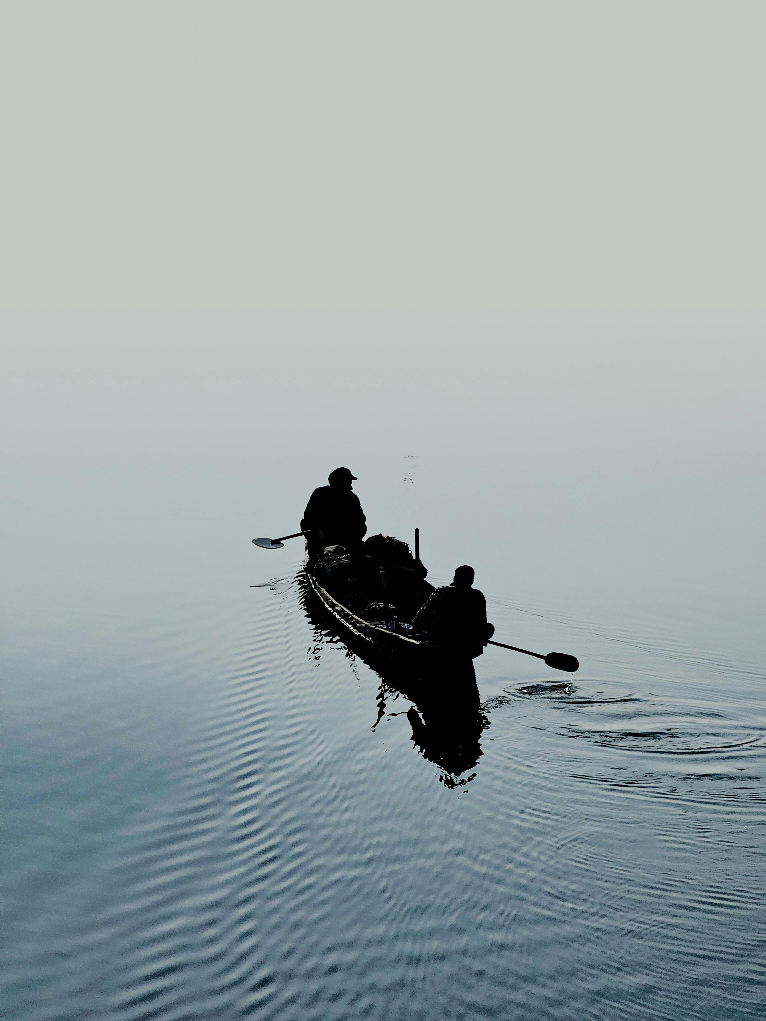 Kostenlos Die Silhouetten zweier Menschen, die im Morgengrauen auf einem ruhigen, nebligen See Kanu fahren, fangen einen friedlichen Moment ein. Stock-Foto