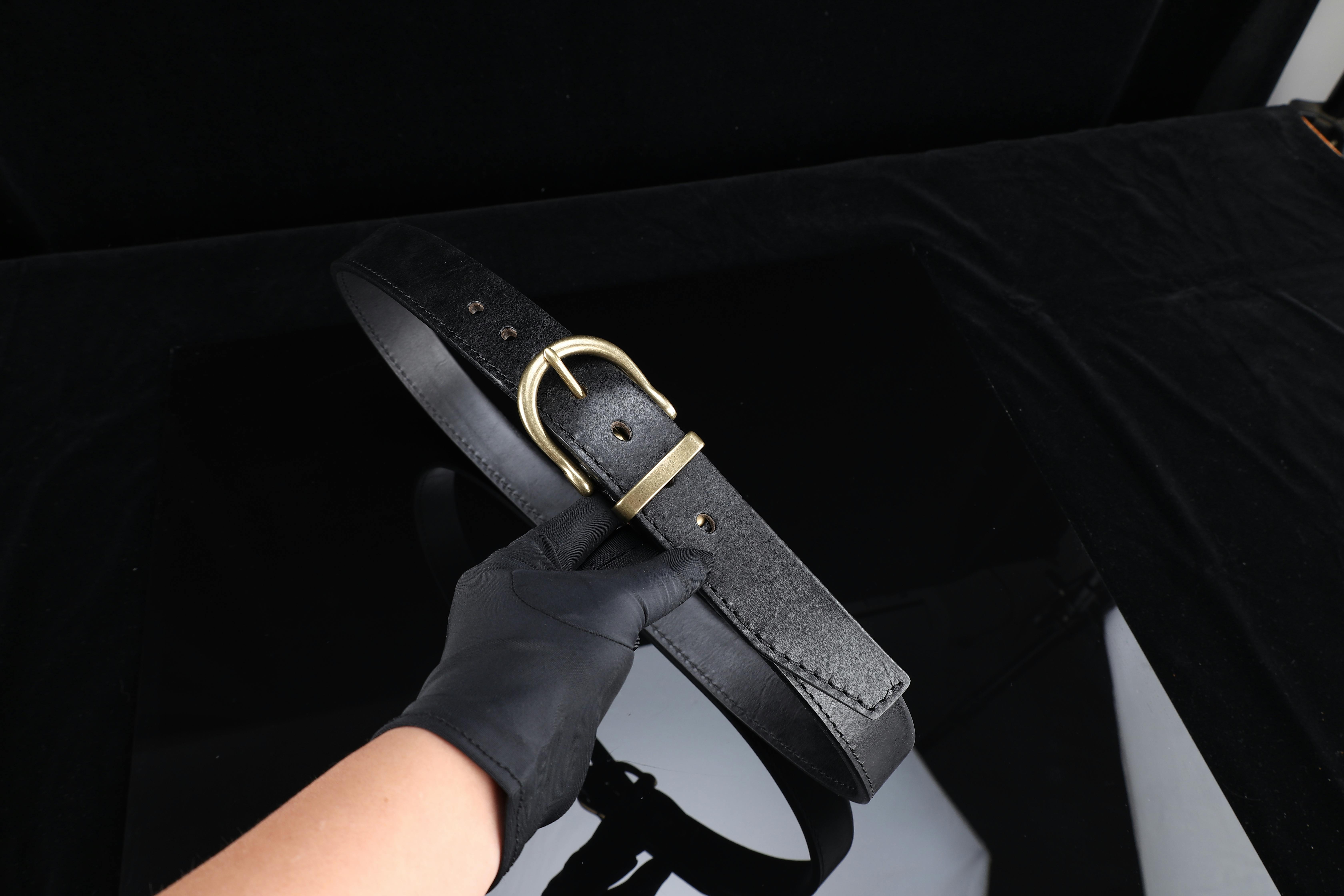 Kostnadsfria Svart läderbälte med ett guldfärgat spänne som hålls av en behandskad hand mot en mörk bakgrund. Stock foto