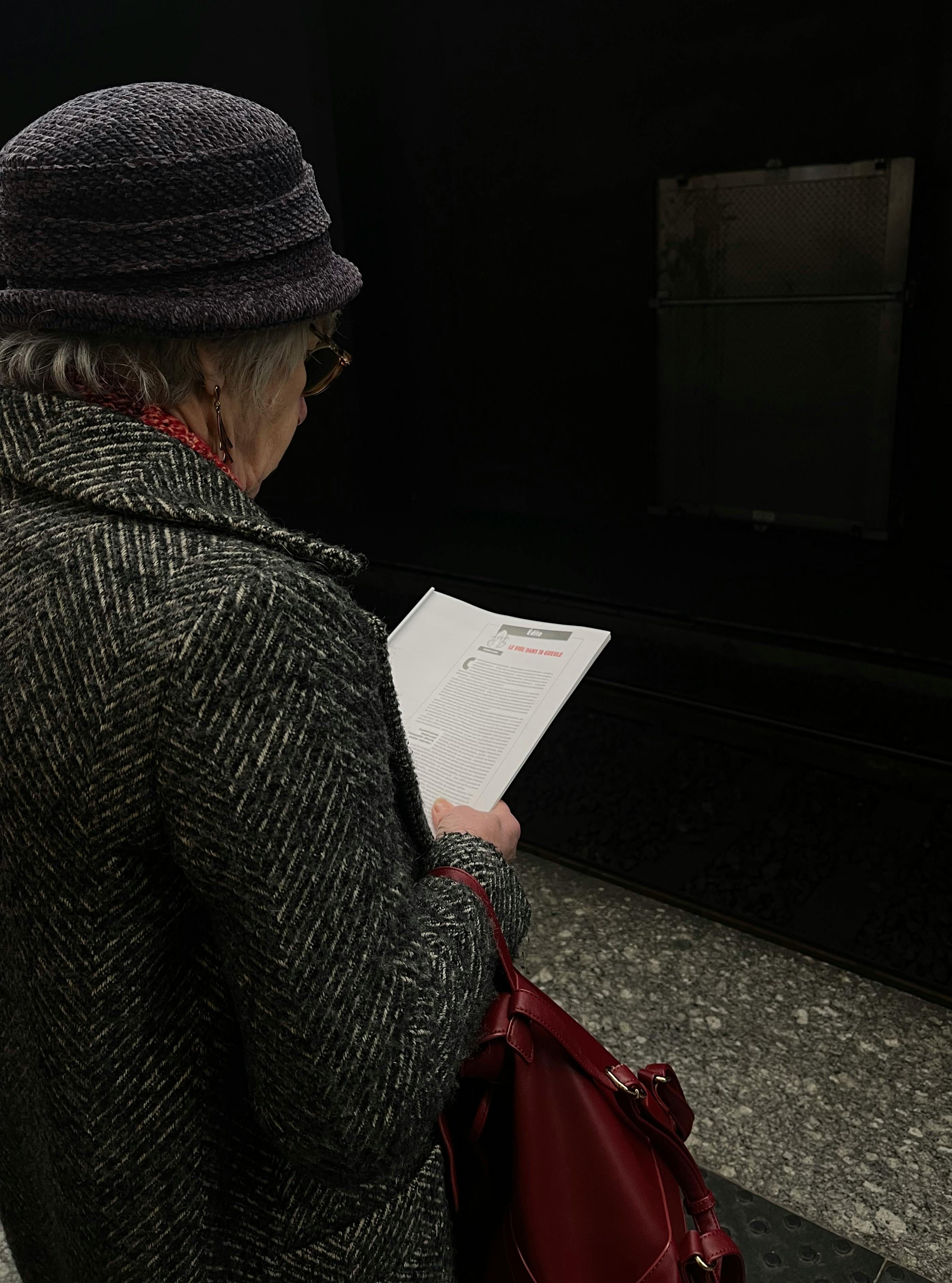 Gratis Eldre kvinne med hatt leser på en svakt opplyst T-baneplattform med en rød veske. Arkivbilde