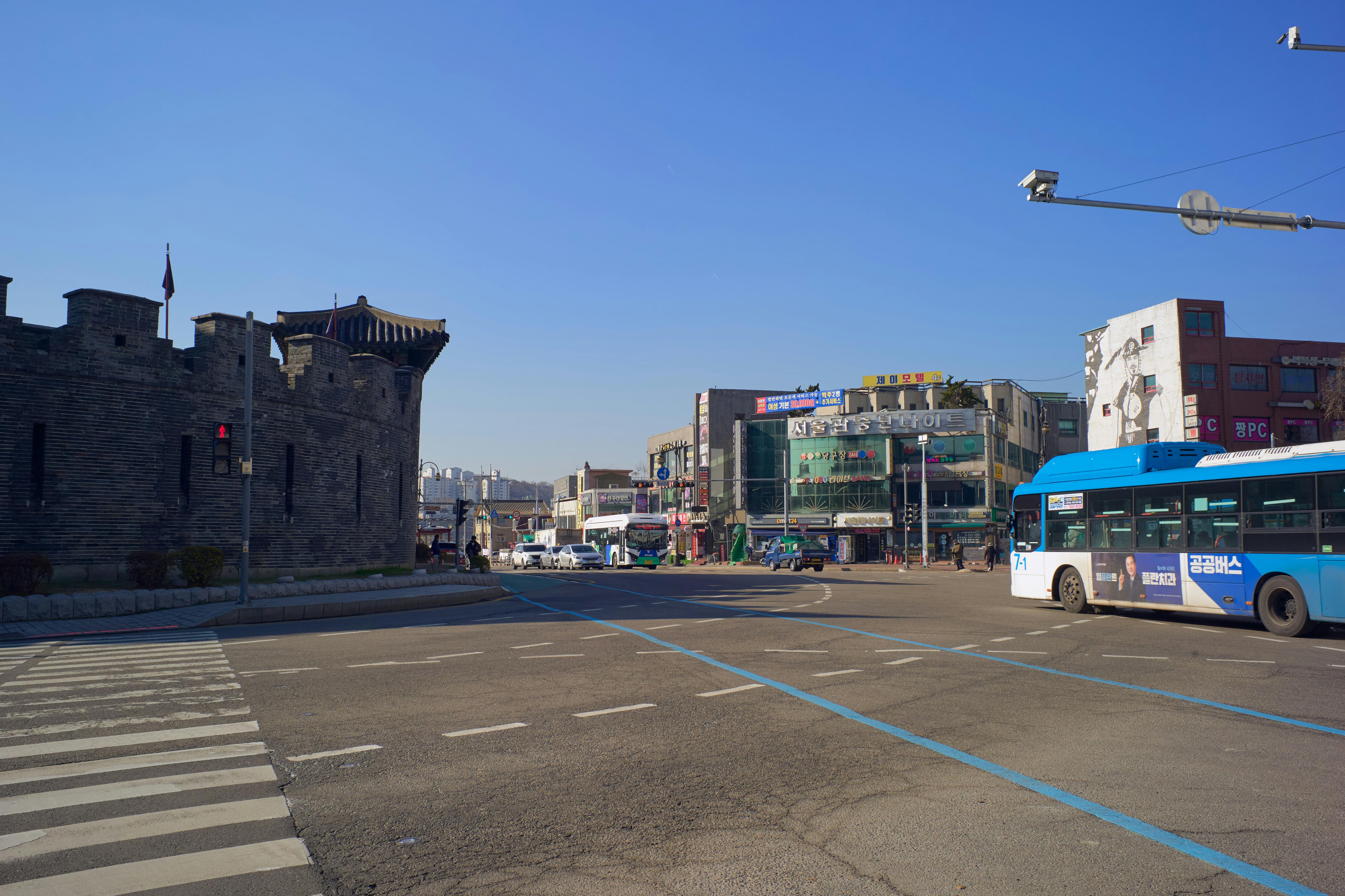 A bustling intersection in front of a historic fortress with buses and shops in broad daylight.