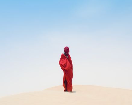 A mysterious figure in a red cloak stands alone on a sand dune under a clear blue sky.