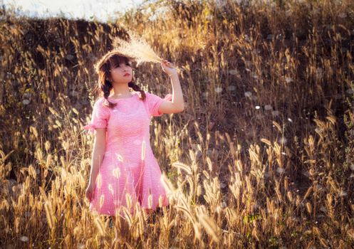A young woman in a pink dress stands in a field of tall grass, backlit by sunlight.