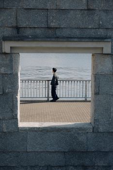 A person stands alone by the water, framed by a stone wall window.