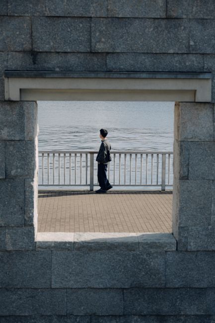 A person stands alone by the water, framed by a stone wall window.