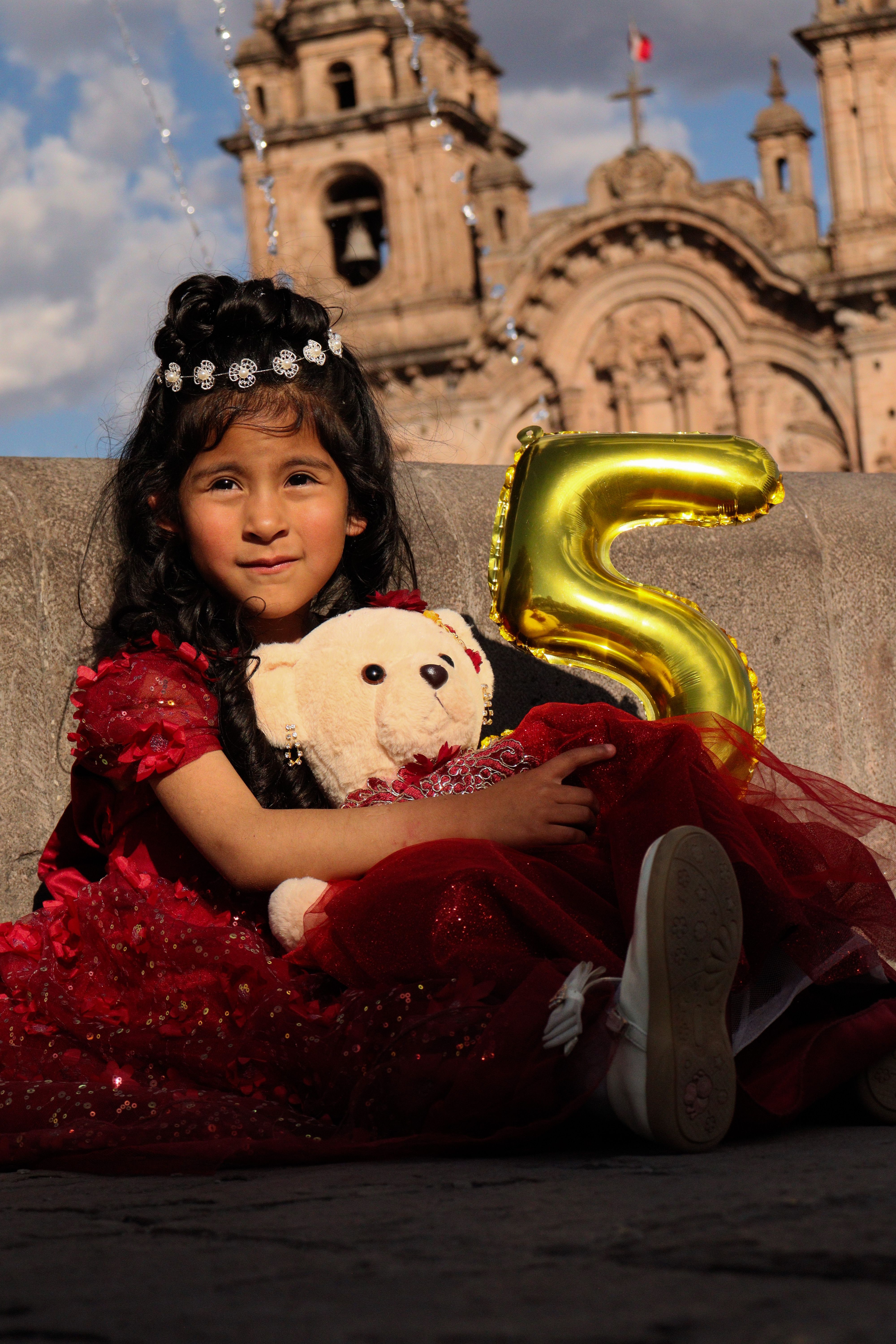Young girl in a red dress holding a teddy bear and a balloon in front of a historic church.