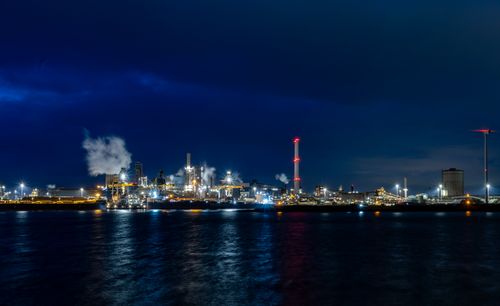 Free A vibrant industrial skyline illuminated against the night sky, reflecting on water. Stock Photo