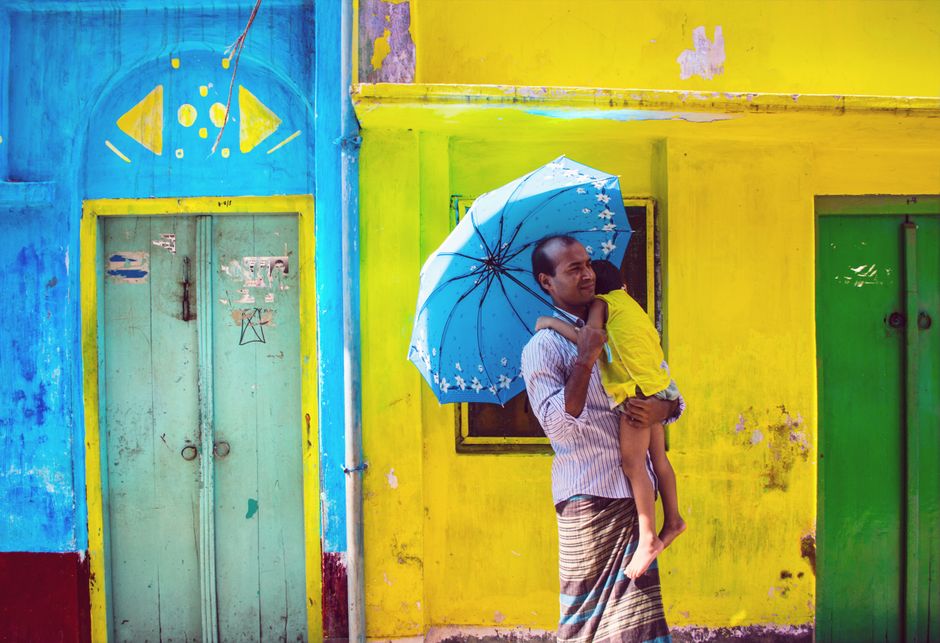 A father holding his child under a blue umbrella stands in front of a colorful, vibrant urban background.