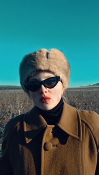 Stylish woman in fur hat and sunglasses in a field under a clear sky.