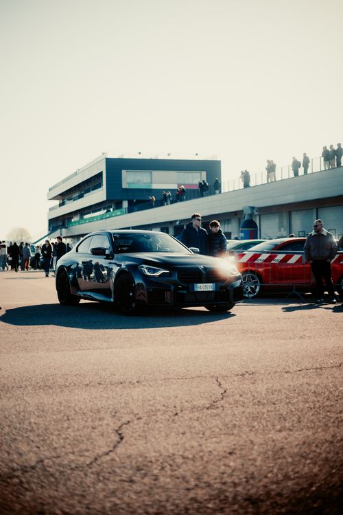 Free A sleek black sports car showcased at a sunny race track gathering with onlookers and modern architecture. Stock Photo