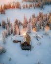 日落时分，从空中俯瞰，一座白雪皑皑的小屋令人惊叹，四周环绕着高大的常绿树木