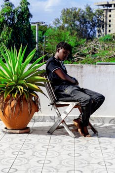 A young adult sits pensively on a patio chair beside a large potted plant.