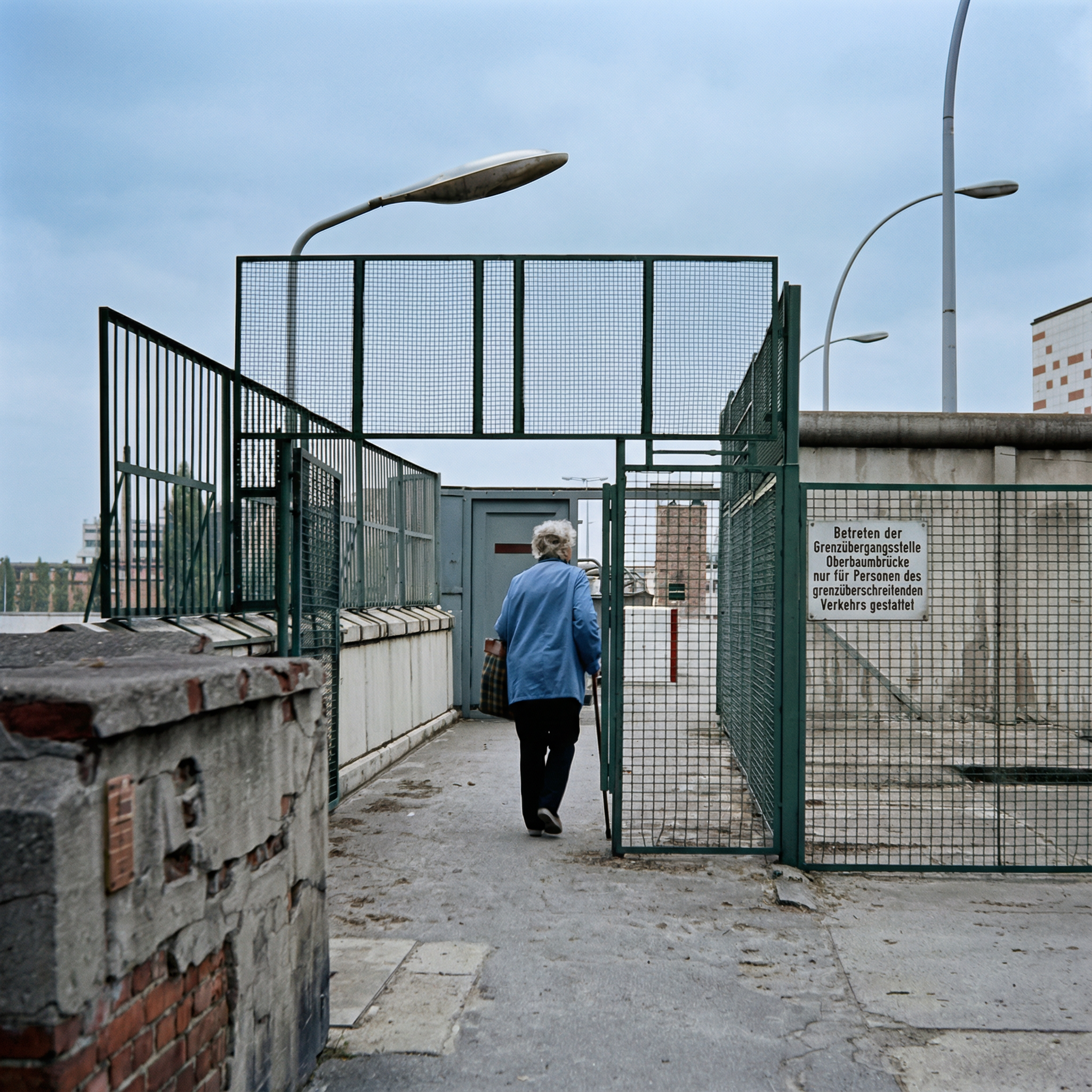 An elderly woman walks alone through a secure gate, suggesting historical or urban exploration.