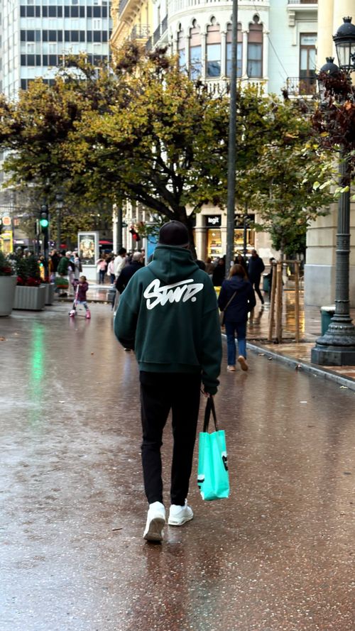 Free Casual stroll during rain in urban cityscape. Man in green hoodie holding turquoise shopping bag. Stock Photo